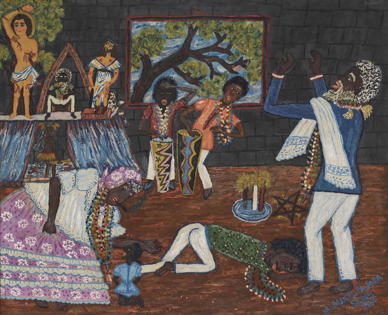Num cômodo de paredes escuras, pessoas com colares participam de uma cerimônia diante de um altar; dois músicos tocam tambores e uma criança fica junto à mulher de saia lilás.
