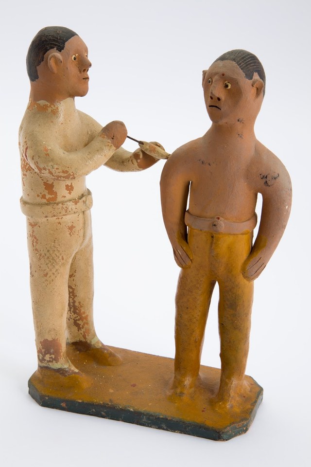 Duas figuras de barro pintado estão de pé numa base estreita; a da esquerda veste branco e encosta um instrumento fino no braço da outra, que tem o torso descoberto e calças amarelas.